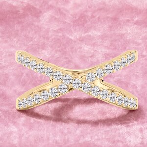 Op de afbeelding: Een goudkleurige ring met een kruisontwerp, bezet met talrijke kleine, sprankelende witte edelstenen. De ring is geplaatst tegen een zachte, roze achtergrond, wat de elegante en delicate uitstraling benadrukt.