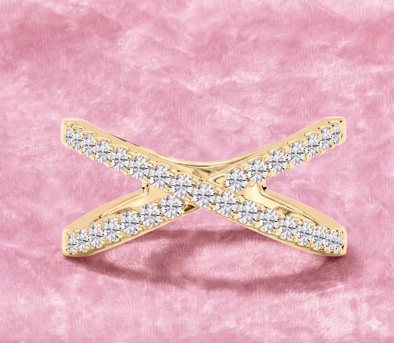 Op de afbeelding: Een goudkleurige ring met een kruisontwerp, bezet met talrijke kleine, sprankelende witte edelstenen. De ring is geplaatst tegen een zachte, roze achtergrond, wat de elegante en delicate uitstraling benadrukt.