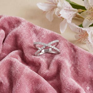 Op de afbeelding: Een zilveren ring met een kruisontwerp, bezet met kleine, sprankelende diamanten. De ring ligt op een zachtroze fluwelen oppervlak, met witte en roze gevlekte bloemen op de achtergrond. Het ontwerp van de ring is modern en elegant.