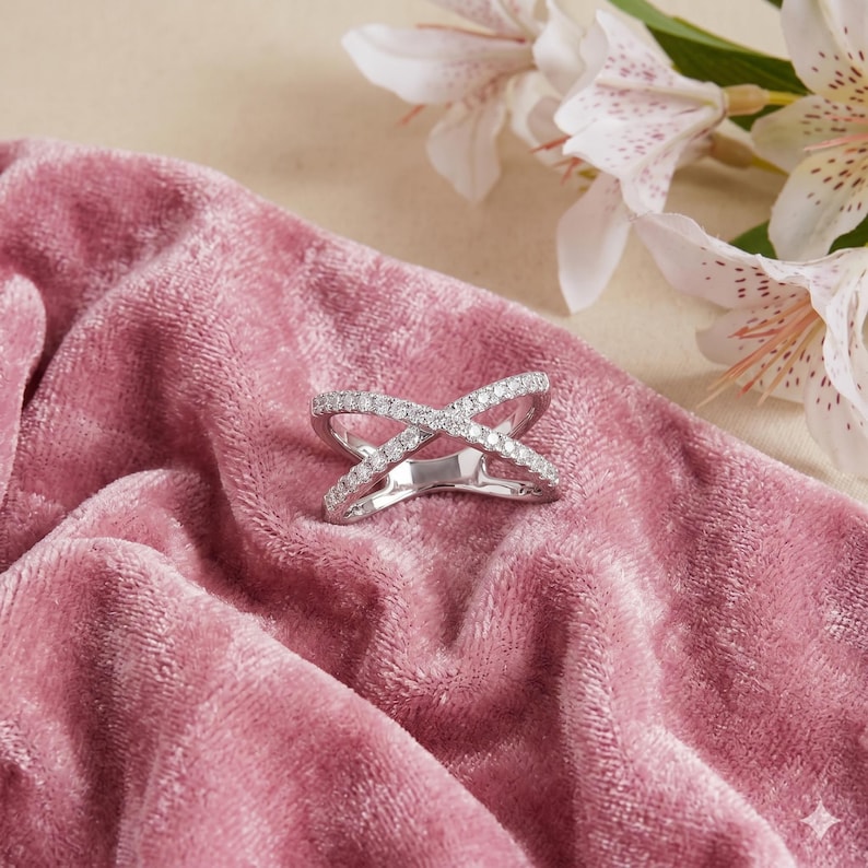 Op de afbeelding: Een zilveren ring met een kruisontwerp, bezet met kleine, sprankelende diamanten. De ring ligt op een zachtroze fluwelen oppervlak, met witte en roze gevlekte bloemen op de achtergrond. Het ontwerp van de ring is modern en elegant.