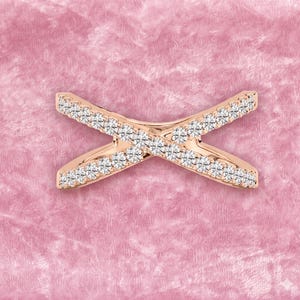 Op de afbeelding: Ros&eacute;gouden ring in X-vorm, bezet met kleine, heldere edelstenen. De ring is geplaatst tegen een zachte roze achtergrond, die de glinstering van de stenen benadrukt. Het ontwerp is symmetrisch en modern, geschikt voor dagelijks gebruik of speciale gelegenheden.