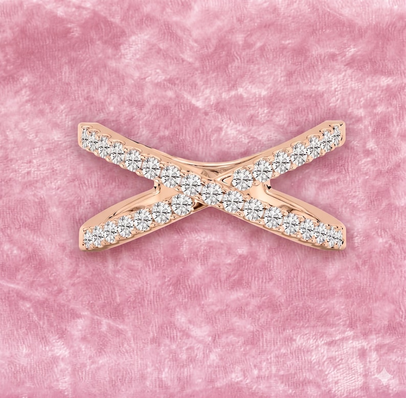 Op de afbeelding: Ros&eacute;gouden ring in X-vorm, bezet met kleine, heldere edelstenen. De ring is geplaatst tegen een zachte roze achtergrond, die de glinstering van de stenen benadrukt. Het ontwerp is symmetrisch en modern, geschikt voor dagelijks gebruik of speciale gelegenheden.
