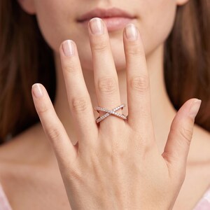 Op de afbeelding: Ros&eacute;gouden kruisring met kleine, heldere edelstenen. De ring wordt om een vinger gedragen en toont zijn elegante ontwerp. De edelstenen geven de sieraden glans.