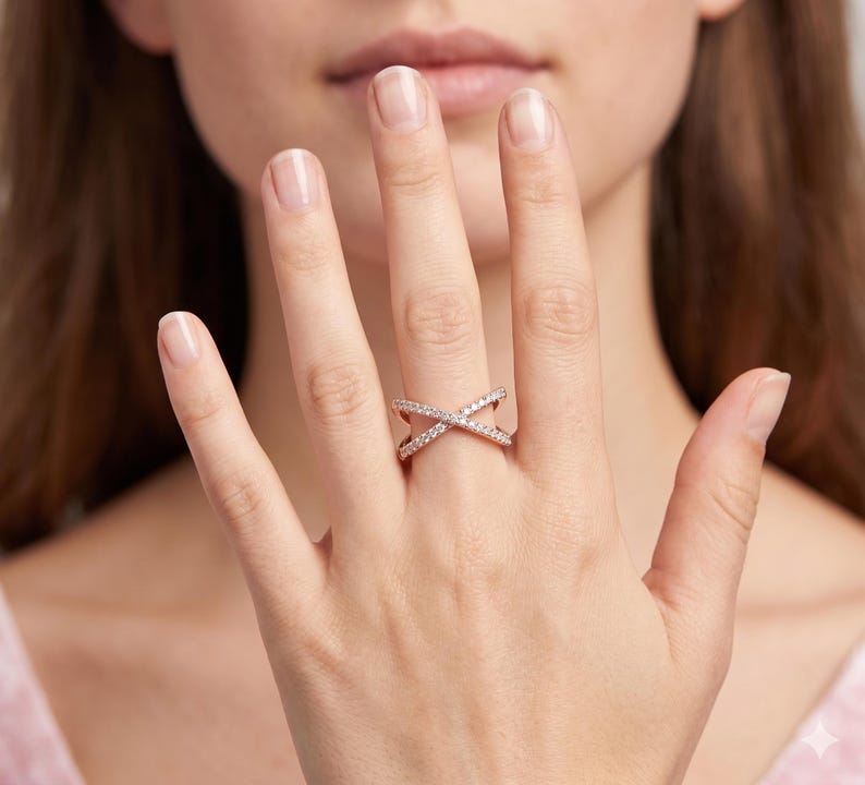 Op de afbeelding: Ros&eacute;gouden kruisring met kleine, heldere edelstenen. De ring wordt om een vinger gedragen en toont zijn elegante ontwerp. De edelstenen geven de sieraden glans.