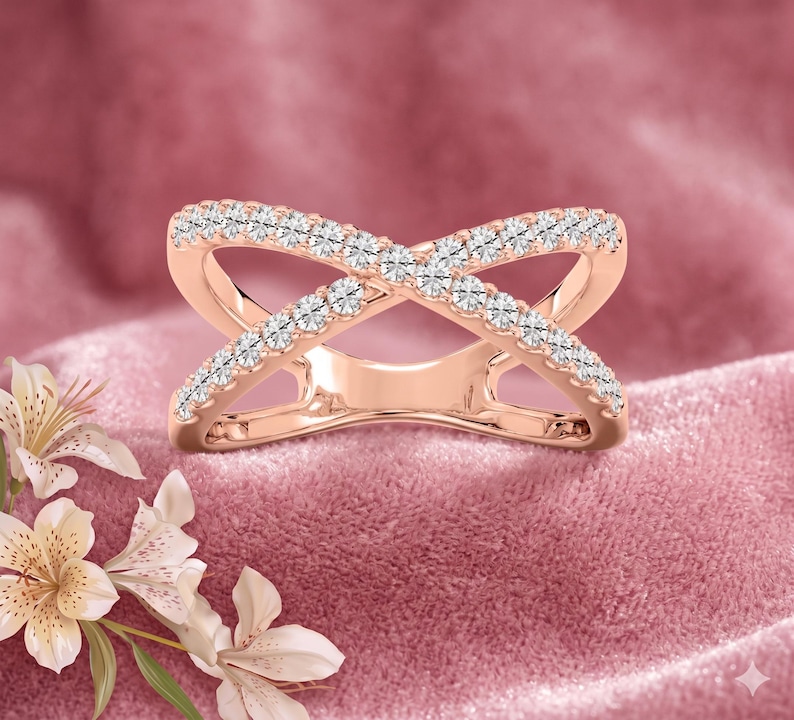 Op de afbeelding: Rosegouden kruisring met een rij kleine, heldere edelstenen langs elke band. De ring is geplaatst tegen een roze achtergrond met witte bloemen in de linkerbenedenhoek. Het ontwerp van de ring vormt een X.