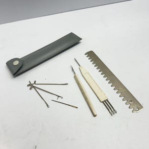 May include: A set of knitting and sewing tools including a grey faux leather case with a snap closure, a long metal serrated gauge, two white plastic-handled latch hooks, a small metal needle, and a multi-pronged metal stitch holder tool.