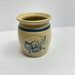 Pode incluir: Um pote de mostarda de grés bege com uma borda larga e corpo cilíndrico. O pote apresenta uma faixa azul horizontal perto do topo e um desenho floral azul abaixo do texto em relevo que diz MUSTARD POT em uma fonte sem serifa clara.
