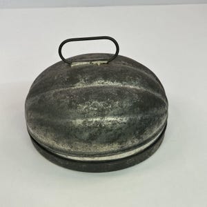 May include: A vintage oval-shaped metal mold with a weathered, dark grey patina and a smooth, rounded top. The mold features a single, thin, dark metal wire handle attached to the center of the lid, designed for lifting the domed cover.
