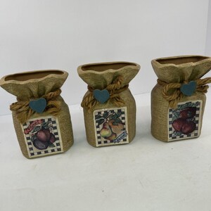 May include: Three tan ceramic vases shaped like tied sacks with a textured finish. Each vase features a central square label with a checkered border displaying illustrations of plums, pears, or apples, accented by a jute rope and a blue heart.