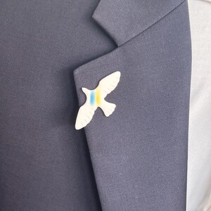 May include: A white ceramic dove pin with blue and yellow stripes on the wings. The pin is worn on a dark blue suit jacket.