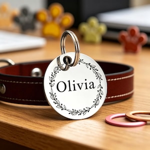 May include: A circular pet tag with the name "Olivia" in black script, framed by a black floral wreath. The tag is attached to a silver ring, next to a brown leather dog collar. Decorative paw-shaped items are in the background.