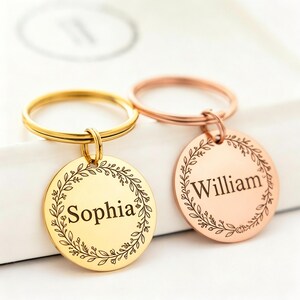 May include: Two personalized keychains, one gold and one rose gold, each with a circular pendant. The gold keychain is engraved with the name "Sophia", and the rose gold keychain is engraved with the name "William". Both pendants feature a floral wreath design.