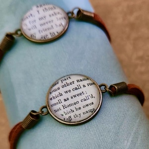 May include: Three brown cord bracelets with circular, antique-style pendants. Each pendant features text from a book, with a bronze-colored metal frame and coiled accents. The bracelets are displayed on a light blue fabric.