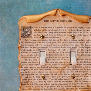 May include: Decorative light switch cover featuring a vintage design with text from "The Little Mermaid." The cover has a gold-colored frame and two white toggle switches. The background is a light blue.