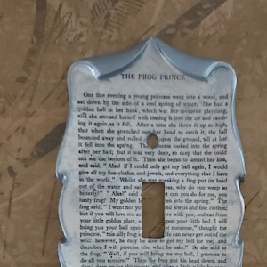 Puede incluir: Cubierta de interruptor de luz decorativa con acabado plateado y un recorte para un interruptor. El diseño vintage incorpora texto de "El Príncipe Rana", añadiendo un toque clásico.