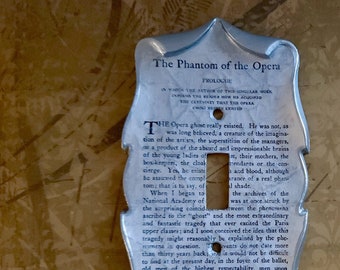 May include: A decorative light switch cover with a silver finish. The cover features the text "The Phantom of the Opera" and excerpts from the story. The design has a shield-like shape.