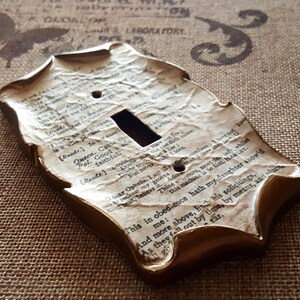 May include: A decorative light switch cover with a vintage aesthetic. The cover is shaped with ornate edges and features a textured surface resembling aged paper with printed text. The edges are painted in a gold color.
