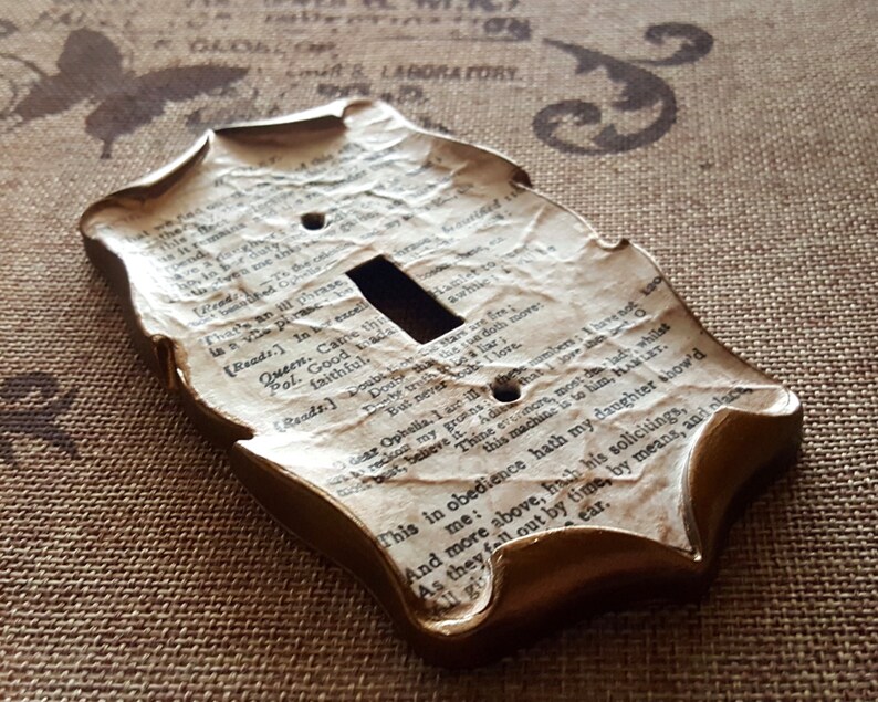 May include: A decorative light switch cover with a vintage aesthetic. The cover is shaped with ornate edges and features a textured surface resembling aged paper with printed text. The edges are painted in a gold color.