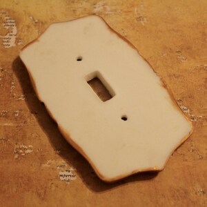 May include: Cream-colored ceramic light switch cover with a rectangular opening and two screw holes. The edges have a rustic, uneven shape with a brown trim. A decorative home decor item.