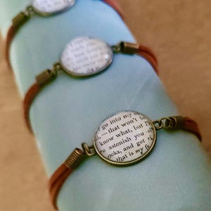 Può includere: Braccialetti in pelle marrone con ciondoli rotondi color bronzo antico. Ogni ciondolo presenta un design testuale stampato sotto una cupola di vetro. I braccialetti sono esposti su una superficie azzurra.