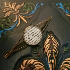 May include: A gold-toned hair clip with a round, white, glass charm featuring a quote from Shakespeare's Romeo and Juliet. The quote reads: "tongue's utters thou not Romeo and Rom. Neither, fair mais Jul. How cam'st thou bit orchard walls are high 't the place death, const of my kinsmen fir With love's"