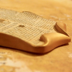 May include: A handmade, decorative piece featuring a section of aged book pages with visible text, set within a curved, gold-painted frame. The frame's edges are rounded, creating a soft, elegant aesthetic. The piece rests on a light brown surface.