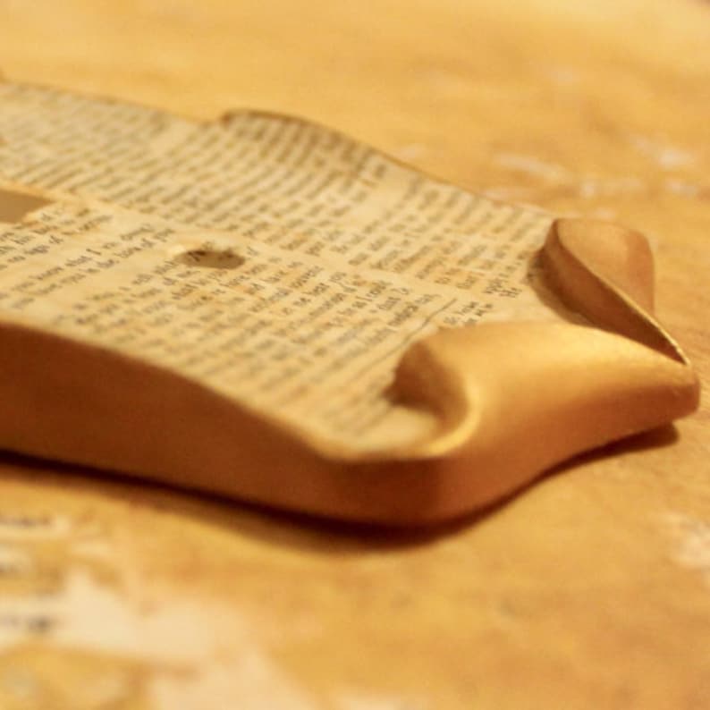 May include: A handmade, decorative piece featuring a section of aged book pages with visible text, set within a curved, gold-painted frame. The frame's edges are rounded, creating a soft, elegant aesthetic. The piece rests on a light brown surface.