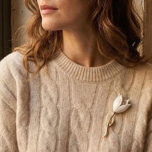 May include: A woman wearing a cream cable-knit jumper with a white tulip brooch. The brooch has a gold stem and is pinned on the left side. The woman is looking out of a window.