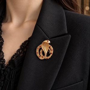 Puede incluir: Un broche de cobra en tono dorado con un cuerpo texturizado y enroscado y una capucha elevada. El diseño de serpiente está acentuado con pequeñas piedras redondas de color rojo vibrante incrustadas a lo largo del cuerpo y un ojo rojo.