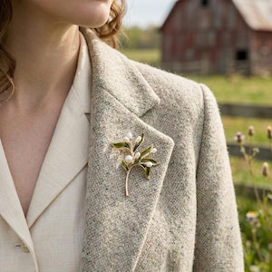 Könnte beinhalten: Goldfarbene Brosche in Form eines Mistelzweigs mit grünen Emailleblättern und weißen, perlenartigen Akzenten. Die Brosche ist an einem grauen Tweed-Blazer befestigt, darunter ein cremefarbenes Hemd. Im Hintergrund ist eine rote Scheune zu sehen.