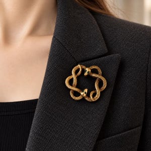 Può includere: Una spilla color oro raffigurante due serpenti intrecciati che formano una figura circolare. I serpenti presentano superfici testurizzate simili a squame e piccole pietre verdi brillanti come occhi, appuntata sul bavero di un blazer grigio scuro.