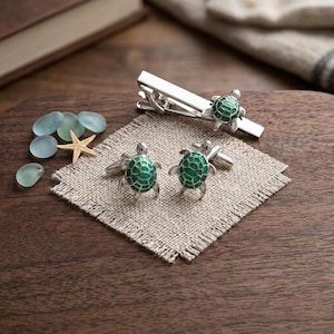 May include: A silver tie clip and a pair of matching cufflinks, each featuring a small sea turtle with a vibrant green enameled shell. The set is displayed alongside several smooth, translucent blue sea glass pebbles and a small beige starfish.