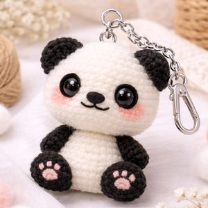 May include: A handmade crochet panda keychain. The panda has a white body, black accents, and pink cheeks. It features a silver-colored chain and clasp. The background includes white fabric, decorative flowers, and small pom-poms.