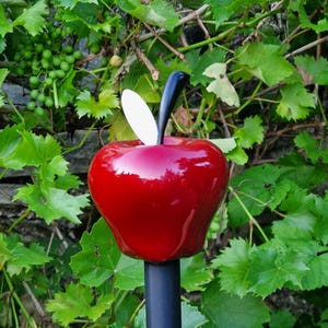 May include: A glossy red apple-shaped garden ornament featuring a smooth, rounded surface, a single white leaf, and a curved black stem. The apple is mounted on a straight, dark gray metal pole, creating a decorative stake for outdoor spaces.