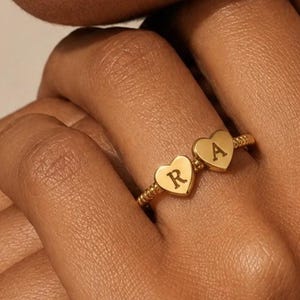 Peut inclure: Une bague dorée ornée de deux breloques en forme de cœur, chacune gravée d'une lettre. La bague a un anneau texturé. Les lettres sont "R" et "A".