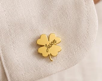 May include: A gold-toned metal cufflink shaped like a four-leaf clover, featuring the name Jason engraved in a cursive script font. The clover-shaped accessory is attached to the cuff of a white, textured fabric shirt with visible stitching.