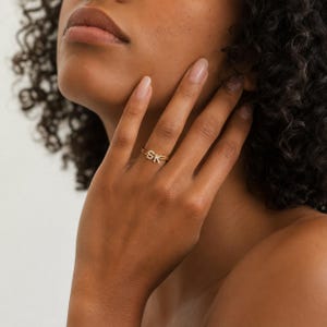 May include: A gold-toned ring featuring the letters S and K as a focal point. The letters are embellished with small, clear, reflective stones. The ring band is thin and metallic, encircling the finger with a simple, polished finish.