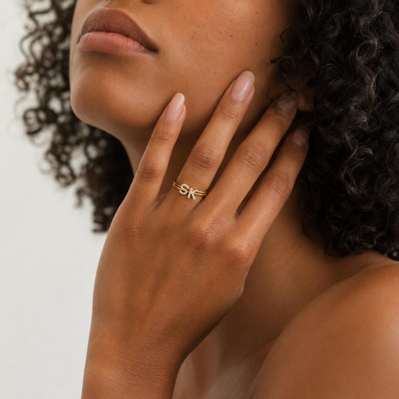 May include: A gold-toned ring featuring the letters S and K as a focal point. The letters are embellished with small, clear, reflective stones. The ring band is thin and metallic, encircling the finger with a simple, polished finish.