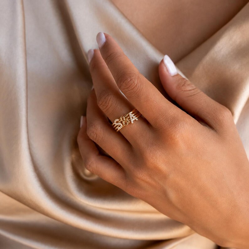 May include: A gold-toned ring set featuring three thin bands, with two bands displaying the letters S and A, and a central band featuring an ampersand symbol. Each letter and the ampersand are embellished with small, clear, sparkling stones.