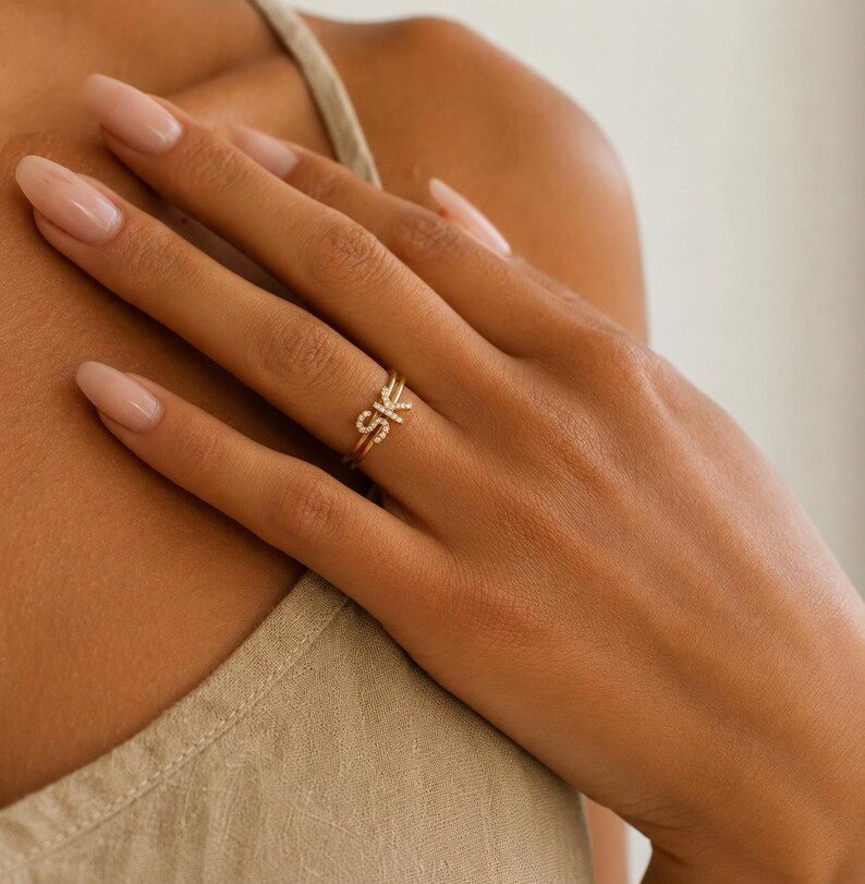 May include: A gold ring featuring the letters S and K, each encrusted with small, clear, sparkling stones. The band is thin and polished, showcasing a delicate design that sits prominently on the finger, highlighting the letter detail.