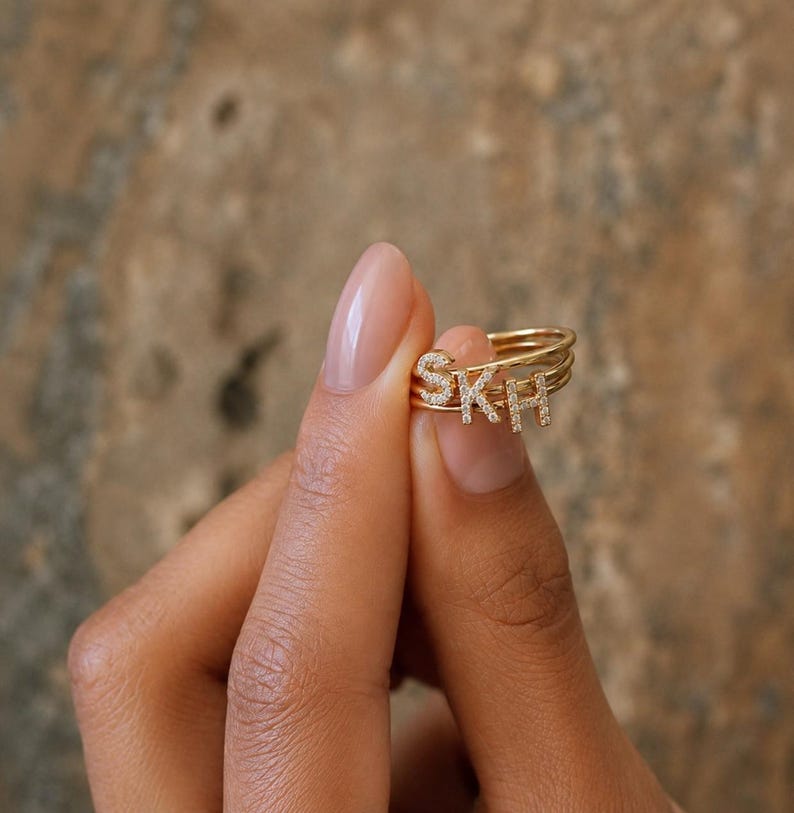 May include: A gold-toned multi-band ring featuring three separate thin bands, each adorned with a single capital letter charm. The letters S, K, and H are encrusted with small, clear, sparkling stones, creating a layered and stacked appearance.