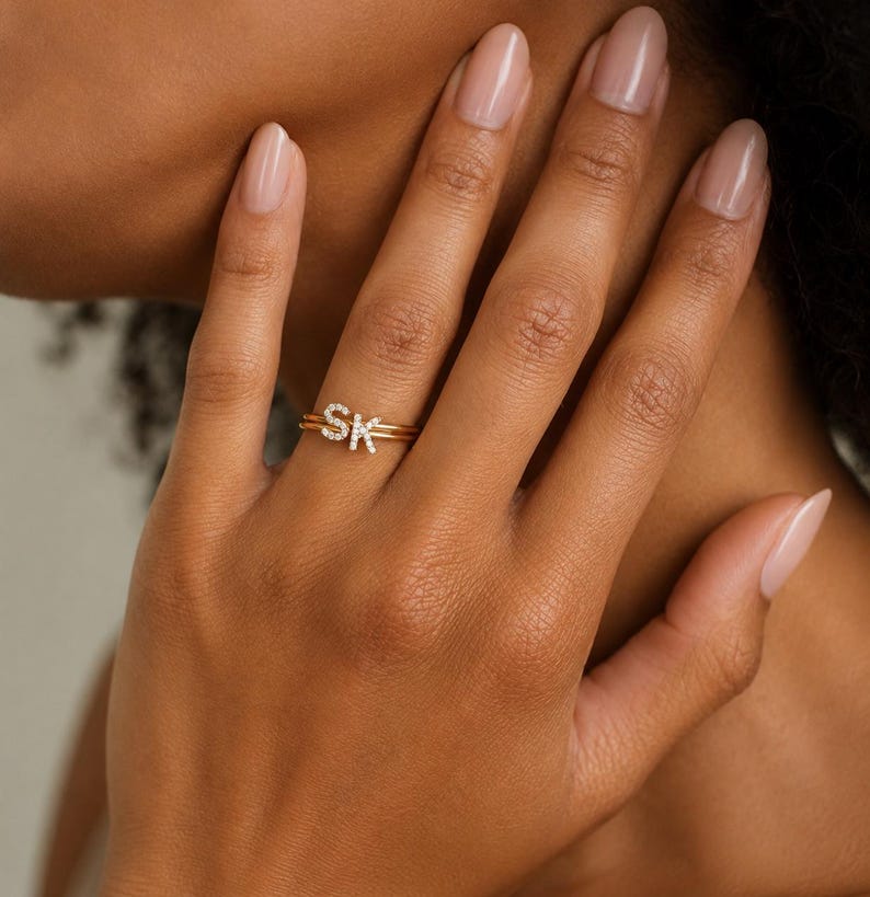 May include: A gold-toned ring featuring two thin bands and two small, capital letter charms, S and K, each encrusted with tiny, clear, sparkling stones. The letters are positioned side-by-side on the top of the ring, creating a personalized design.