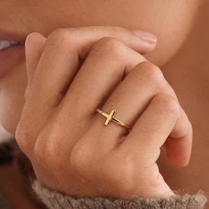 Peut inclure: Une bague en or poli présentant un petit motif de croix centré. L'anneau est fin et lisse, avec une finition métallique réfléchissante qui capte la lumière. La croix est intégrée à l'anneau, créant une esthétique simple et épurée.