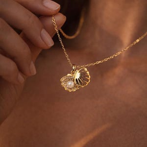 May include: A gold-toned necklace featuring a pendant shaped like an open seashell. The shell contains a single white pearl at its center and is accented with small, clear sparkling stones along the inner edge of the shell's bottom half.