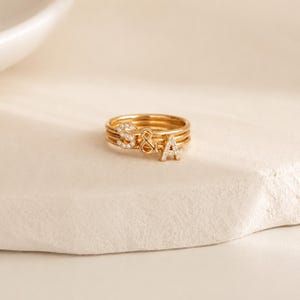 May include: A gold-toned ring set consisting of three thin bands stacked together. The ring features two letter charms, S and A, each encrusted with small clear stones, separated by a small gold-toned ampersand symbol in the center.