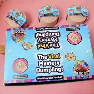 May include: A blue box with the text "The Viral Mystery Dumpling!" and illustrations of dumplings. Three round, wooden containers with colorful labels are also visible. The box reads "What Color Will You Get?"