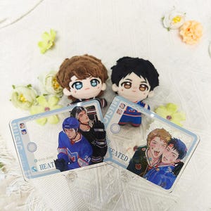 May include: Two plush dolls with brown and black hair, accompanied by two rectangular acrylic stands featuring anime-style illustrations of hockey players and young people. The stands have light blue borders and text including MUSIC and HEATED.
