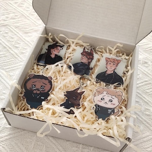 May include: A white cardboard box filled with shredded paper filler containing six acrylic keychains. The keychains feature various illustrated characters, including human figures and stylised animal-like creatures in dark clothing.
