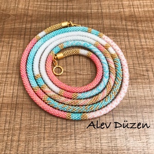 May include: A multi-colored beaded necklace with a gold clasp. The necklace is made of pink, white, and turquoise beads. The necklace is coiled up on a wooden surface.