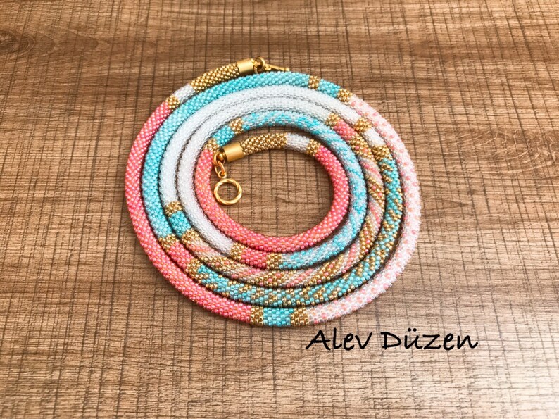 May include: A multi-colored beaded necklace with a gold clasp. The necklace is made of pink, white, and turquoise beads. The necklace is coiled up on a wooden surface.